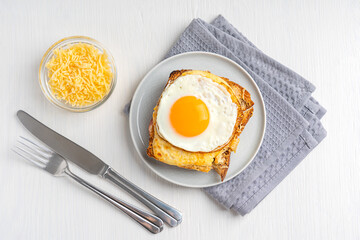 Top view of Croque Madame French baked hot sandwich made of ham and cheese inside toasted bread topped with fried egg with yolk served on plate with tableware and towel on white wooden background