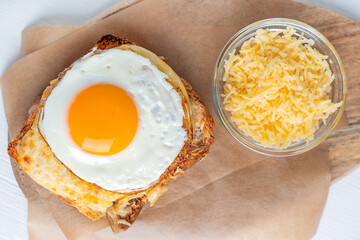 Close up top view of Croque Madame French baked homemade hot sandwich made of ham and cheese inside toasted bread topped with fried egg with yolk served on parchment paper on wooden board on table