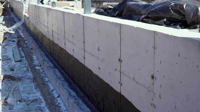 A worker pouring concrete mix formwork on a concrete retaining wall. Construction of a modern wooden house from glued beams. Bituminous membrane waterproofing for concrete foundation.