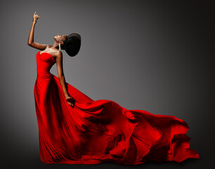 Fashion African Woman in Silk Dress dancing. Dark Skinned Model with Black Afro Hair in Long Evening Red Gown with Tail Fabric flying over Gray Background. Women Luxury Clothing Side View - 499774402