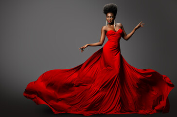 Fashion Model in Red Long waving Luxury Dress. Dark Skinned Beauty Woman with Afro Black Hairstyle dancing over Gray Background. Happy African Sexy Girl in Silk Evening Gown with flying Fabric - Powered by Adobe