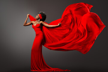 Fashion African Woman in Red Dress with Silk Scarf flying on Wind. Happy Dark Skinned Model Dancing with Fluttering Chiffon Fabric over Gray Background. Afro Female in Long Gown with waving Cloth - 499774415