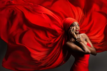 Afrikanische schwarze Frau in rotem Seidenkopftuch. Mode dunkelhäutiges Model-Porträt in Turban mit goldenem Schmuck im Hijab, der Chiffon-Stoff über grauem Hintergrund fliegt. Stilvolle arabische Schönheits-Frauen-Kopfbedeckung © inarik