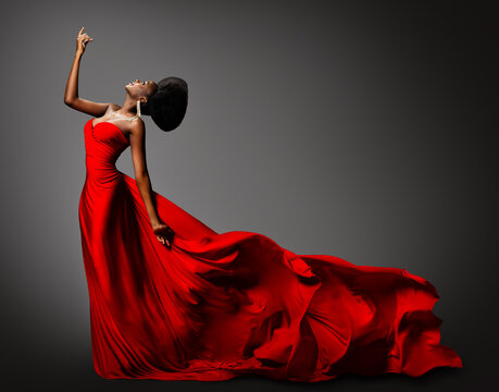 Fashion African Woman in Silk Dress dancing. Dark Skinned Model with Black Afro Hair in Long Evening Red Gown with Tail Fabric flying over Gray Background. Women Luxury Clothing Side View