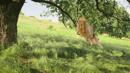 young happy joyful playful blonde woman rides on swing. Hair, long dress fly in wind. Summer spring nature green grass tree. Girl fashion model smiling face. Back rear view. Bridal vintage silk gown