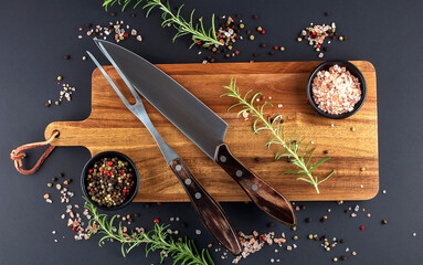 Wooden cutting board, rosemary and spices with fork and knife carving set on dark background