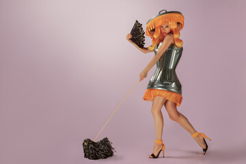 Gorgeous young woman with yellow hair, wearing a fringed trash can as a dress and hat, walking her small black dog made of strips of plastic garbage bag, and holding a fan