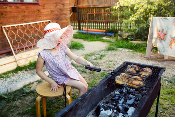 Funny girl kid in a hat at the barbecue grill, meat on the grill and the child is watching the...