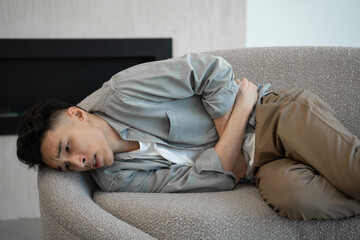 Young Asian man suffers from diarrhea attack putting hand on belly. Exhausted guy lies on sofa...