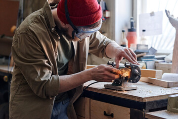 Professional young male carpenter wearing safety glasses working in joinery workshop sharpening knife using grinding jig - obrazy, fototapety, plakaty