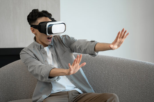 Asian man in goggles enjoys virtual reality getting scared of terrifying images. Young guy in casual clothes sits on sofa at home moving hands