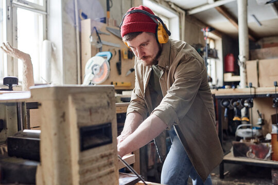 Young male carpenter wearing noise-cancelling headphones using thickness planer machine in woodworking workshop