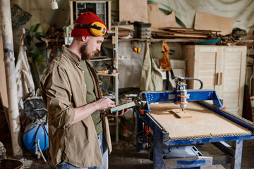 Modern professional carpenter standing in woodworking workshop using digital tablet to do calculations