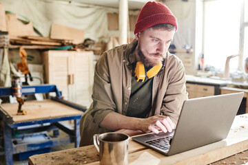 Young bearded carpenter with hearing protection headphones over his neck surfing Internet on laptop...