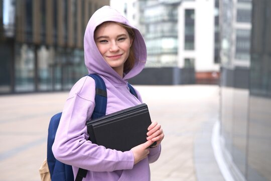 Portrait of happy teen teenager girl, young woman university or college academic student in hoodie with purple hood on her head is holding books, textbooks in hands, looking at camera and smiling