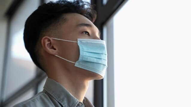 Focused Asian employee wears medical mask on face looking out of window in office. Young man takes protective measures during covid pandemic closeup