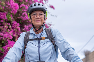 Attractive smiling senior cyclist woman on electric bicycle cycling outdoors wearing helmet passing close to a blooming pink bougainvillea. Elderly woman enjoying healthy lifestyle