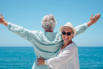 Cheerful senior woman with hat hugging her husband while looks at horizon over sea with outstretched arms. Happy retired couple enjoying summer holidays. Copy space