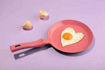 Creative idea with a pink frying pan and egg in heart shape on purple background. Minimal food and love concept. Breakfast idea for Valentine's day and romantic morning.