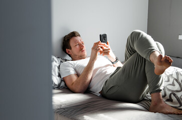 Caucasian attractive young man in white t-shirt woke up chatting, surfing, working on pc and making phone call on bed in bedroom interior. Modern device for remote communication