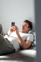Caucasian attractive young man in white t-shirt woke up chatting, surfing, working on pc and making phone call on bed in bedroom interior. Modern device for remote communication