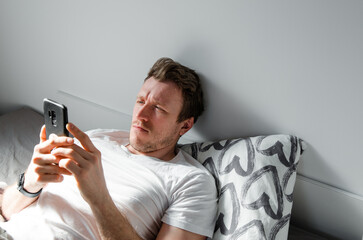 Caucasian attractive young man in white t-shirt woke up chatting, surfing, working on pc and making phone call on bed in bedroom interior. Modern device for remote communication