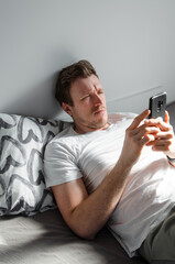 Caucasian attractive young man in white t-shirt woke up chatting, surfing, working on pc and making phone call on bed in bedroom interior. Modern device for remote communication