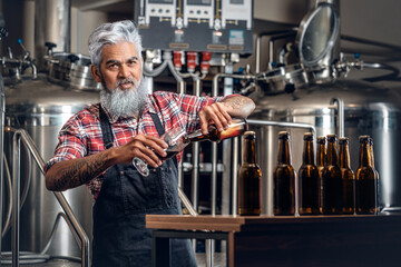Shot of bearded senior brewer pouring his product to glass around table with beer and barrels.