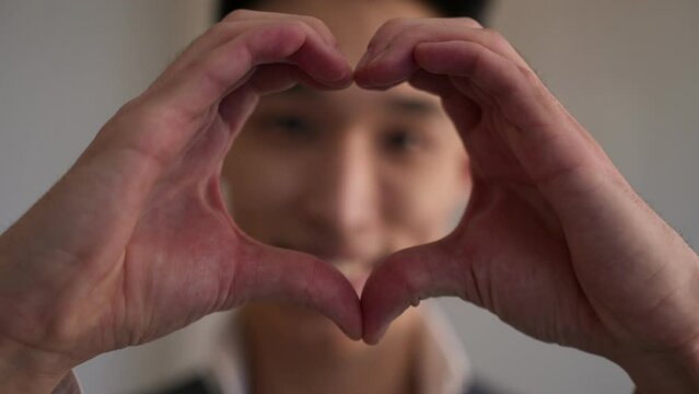 Handsome Asian man shows love gesture smiling and looking in camera. Focus changes from young positive guy to hands making heart shape closeup