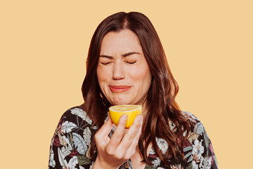 Face portrait of woman wears floral shirt, eating acid lemon doing funny face expression with eyes closed. Studio shot over yellow background.  - obrazy, fototapety, plakaty