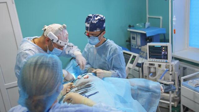 Two operating surgeons assisted by the female nurse. Performing operation in modern equipped surgery room.