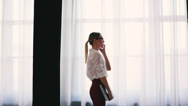 4k Elegant businesswoman in glasses and white shirt. Talking on the phone near big office window.