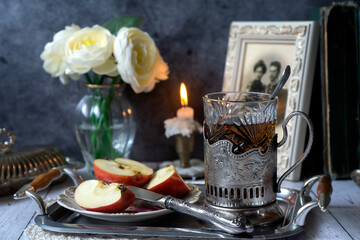 In the foreground is a glass of hot tea in a silver cup holder, sliced apples and other items. Still life in vintage style.
