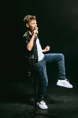 Portrait of Caucasian teenager in white t-shirt, blue jeans and leather jacket with microphone singing on dark background. Hobby and glory concept