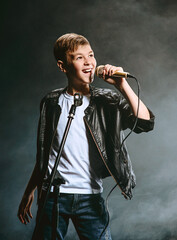 Portrait of Caucasian teenager in white t-shirt, blue jeans and leather jacket with microphone singing on dark background. Hobby and glory concept