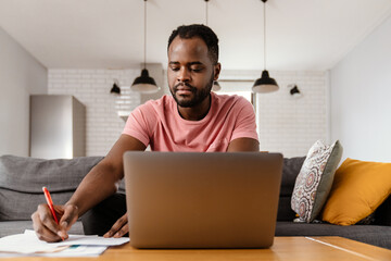 Black man writing down notes while working with laptop and papers