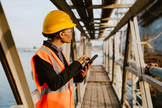 Black Woman Wearing Helmet And Vest Working With Tablet Computer In Port