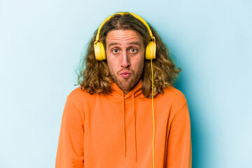 Fototapeta premium Young caucasian man listening to music isolated on blue background shrugs shoulders and open eyes confused.