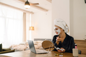 White woman wrapped towel applying face mask while looking in mirror