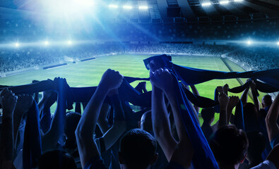 Back view of football, soccer fans cheering their team with scarfs at crowded stadium at evening...