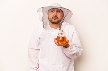 Hispanic beekeeper man holding honey isolated on white background shrugs shoulders and open eyes...