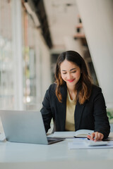 Smiling Asian businesswoman working on paper and holding pen and laptop sitting on a chair at modern home studio. Mobile workers at work and calculators Finance or Accounting Concepts