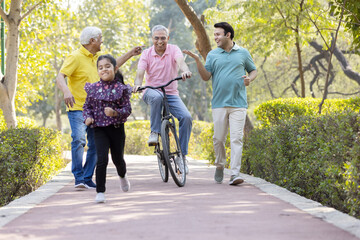 Senior man riding bicycle with granddaughter running while other old man and son encouraging at park