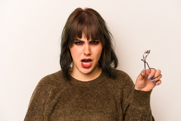 Young caucasian makeup artist woman holding a eyelash curler isolated on white background screaming...