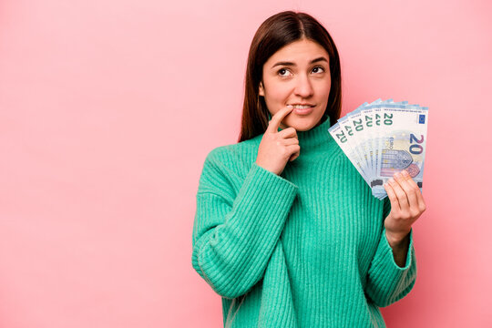 Young caucasian woman holding banknotes isolated on pink background relaxed thinking about something looking at a copy space.