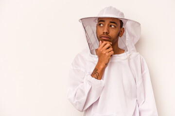 Young African American beekeeper isolated on white background looking sideways with doubtful and skeptical expression.
