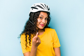 Young hispanic woman wearing a helmet bike isolated on blue background pointing with finger at you as if inviting come closer.