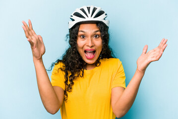 Young hispanic woman wearing a helmet bike isolated on blue background receiving a pleasant surprise, excited and raising hands.