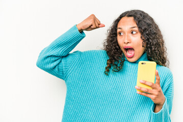 Young hispanic woman holding mobile phone isolated on white background raising fist after a victory, winner concept.