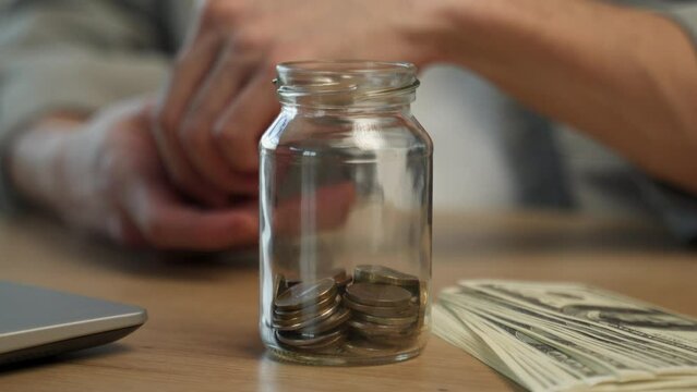 Young employee throws coins in glass jar near dollar bills and laptop at wooden table. Man separates money after getting salary in office closeup